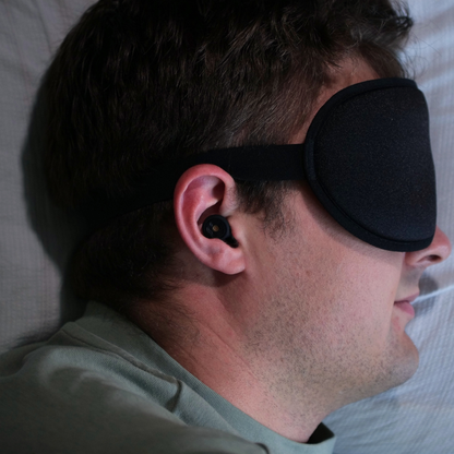Man wearing a black sleep mask and silicone earplugs lying down in a bed.